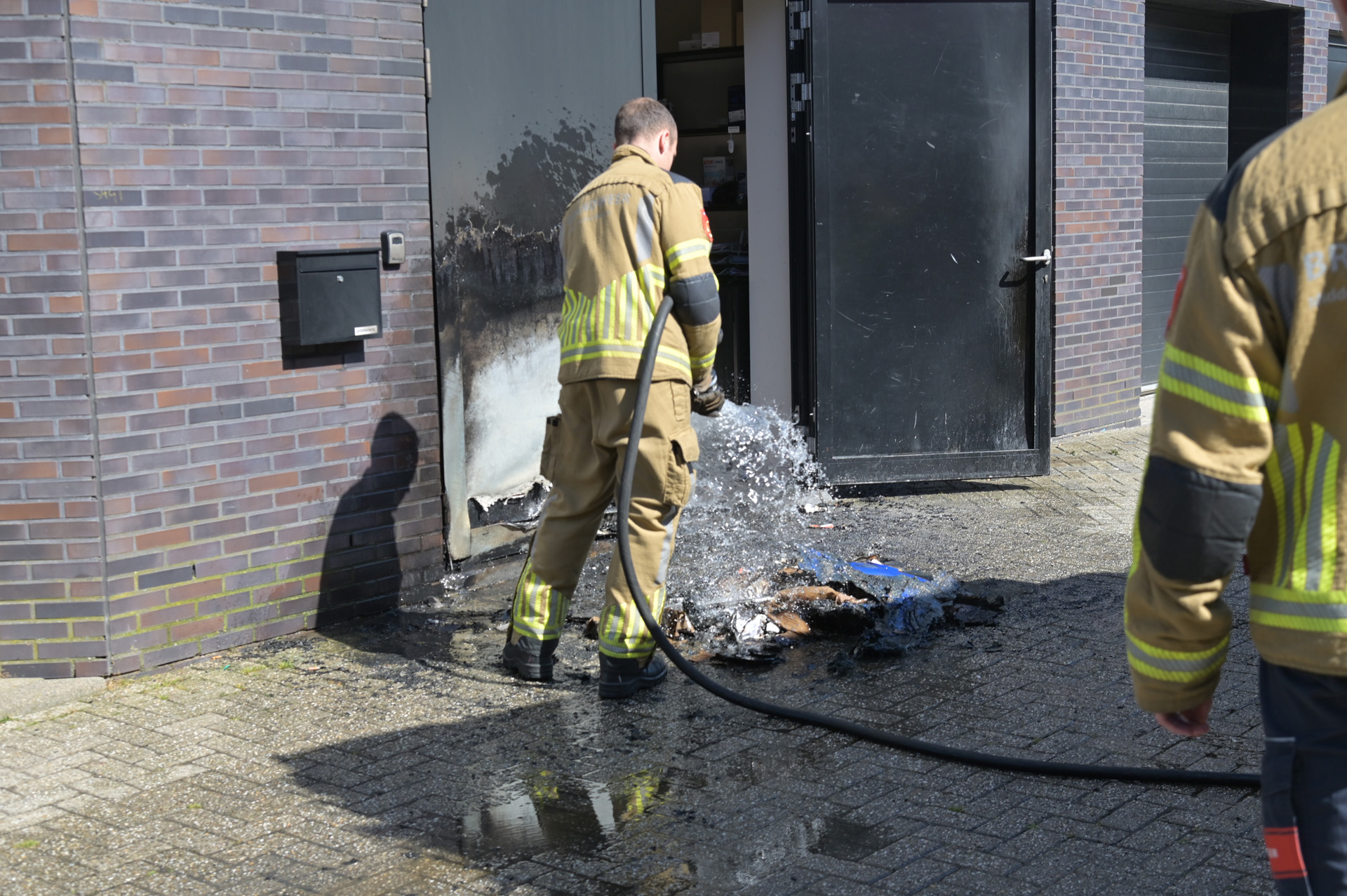 Firefighters hose down a burned doorway outside a brick building, water spraying onto charred debris on the pavement.