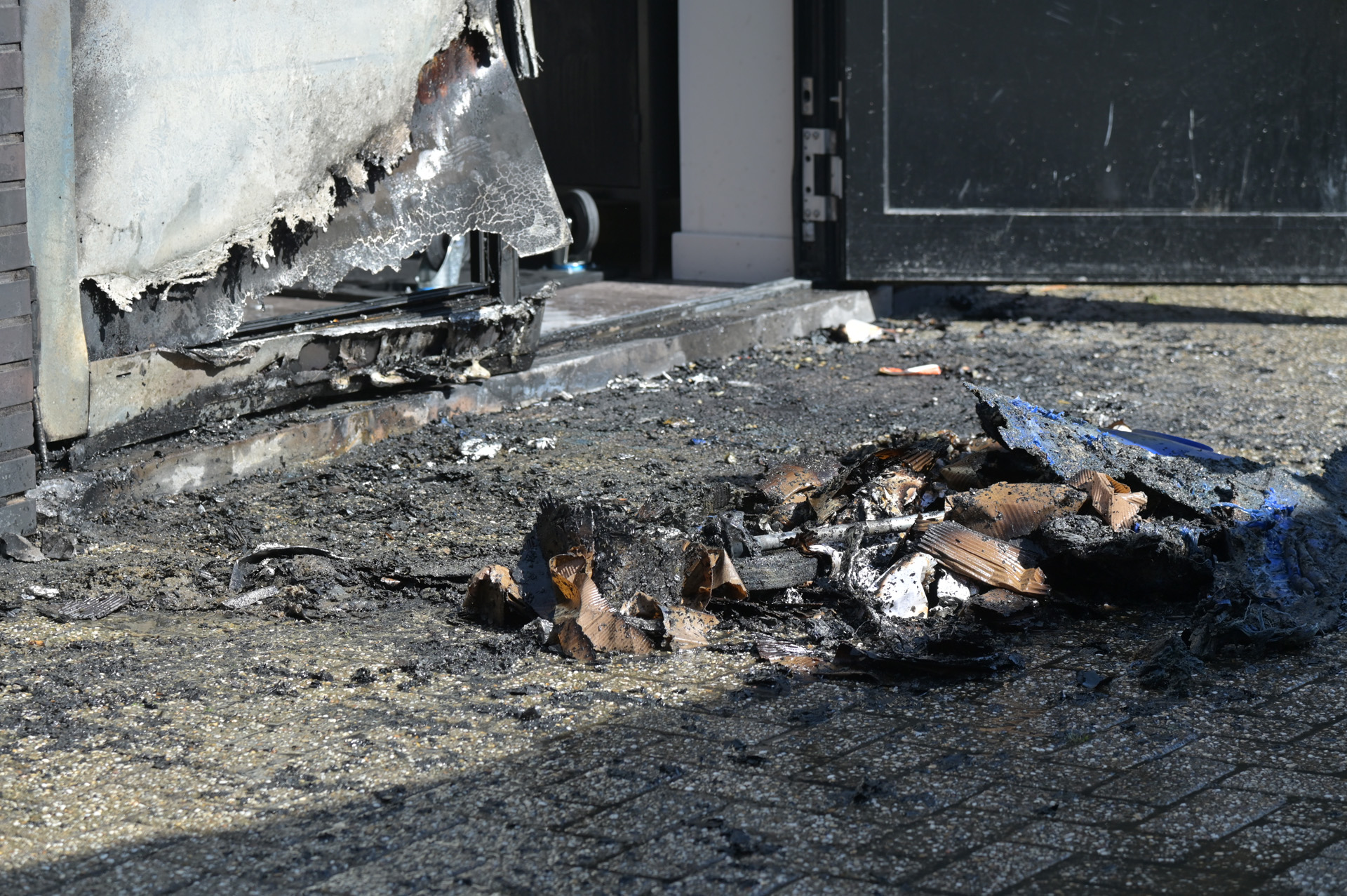 Charred debris and melted insulation along a sidewalk beside a damaged doorway after a fire in a building exterior area.