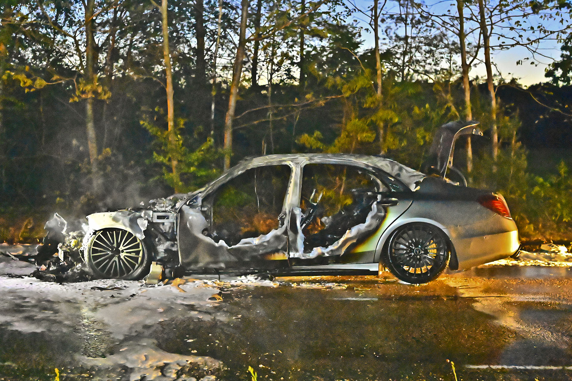 Charred sedan wreck on a road with smoke rising, set against a forest backdrop.