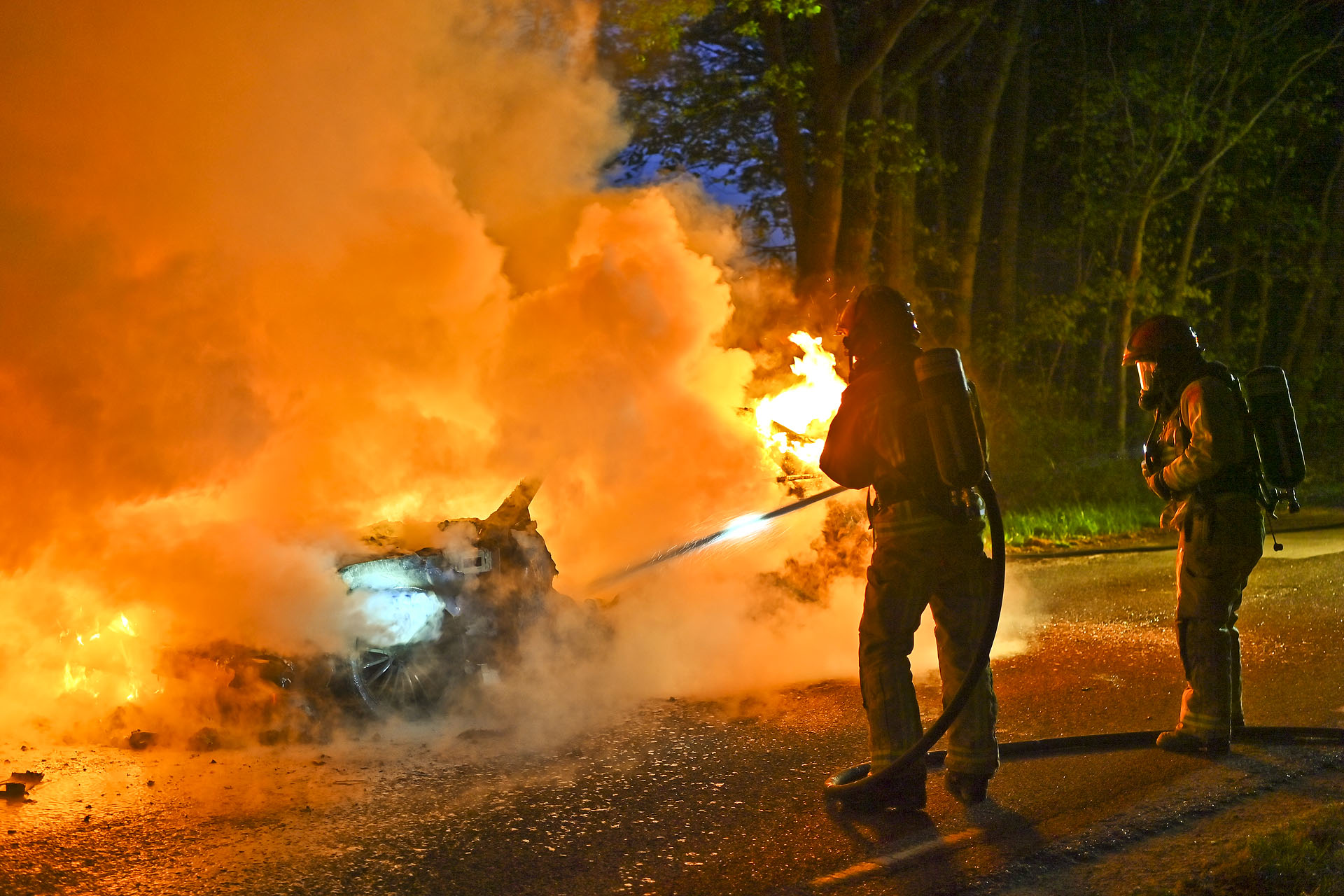 Firefighters in protective gear spray water on a blazing vehicle, thick orange smoke filling the scene at night.