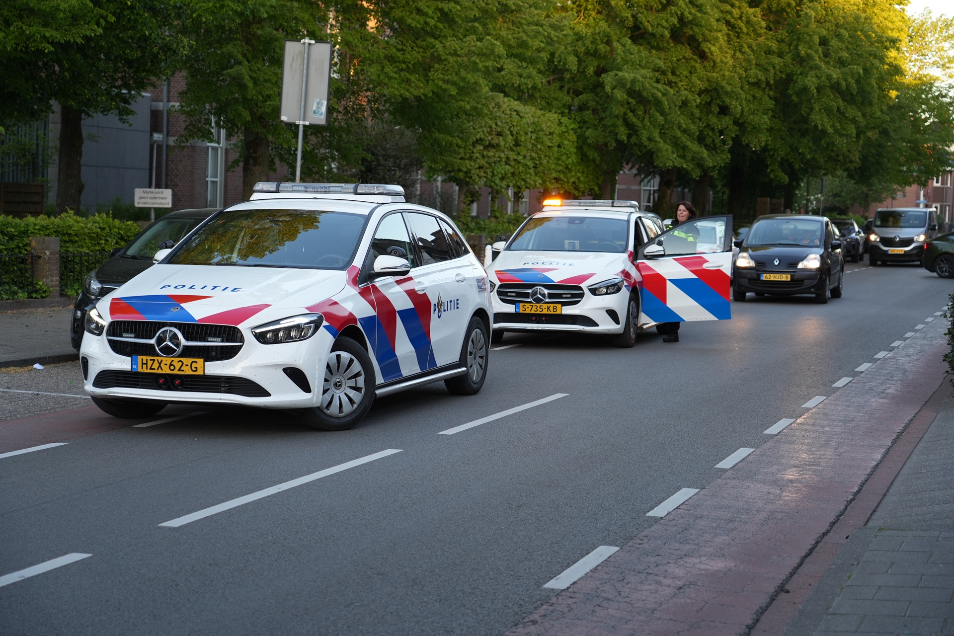 Dutch police cars with blue-red chevrons parked along a residential street; officer stands by open door of a white patrol car with flashing light
