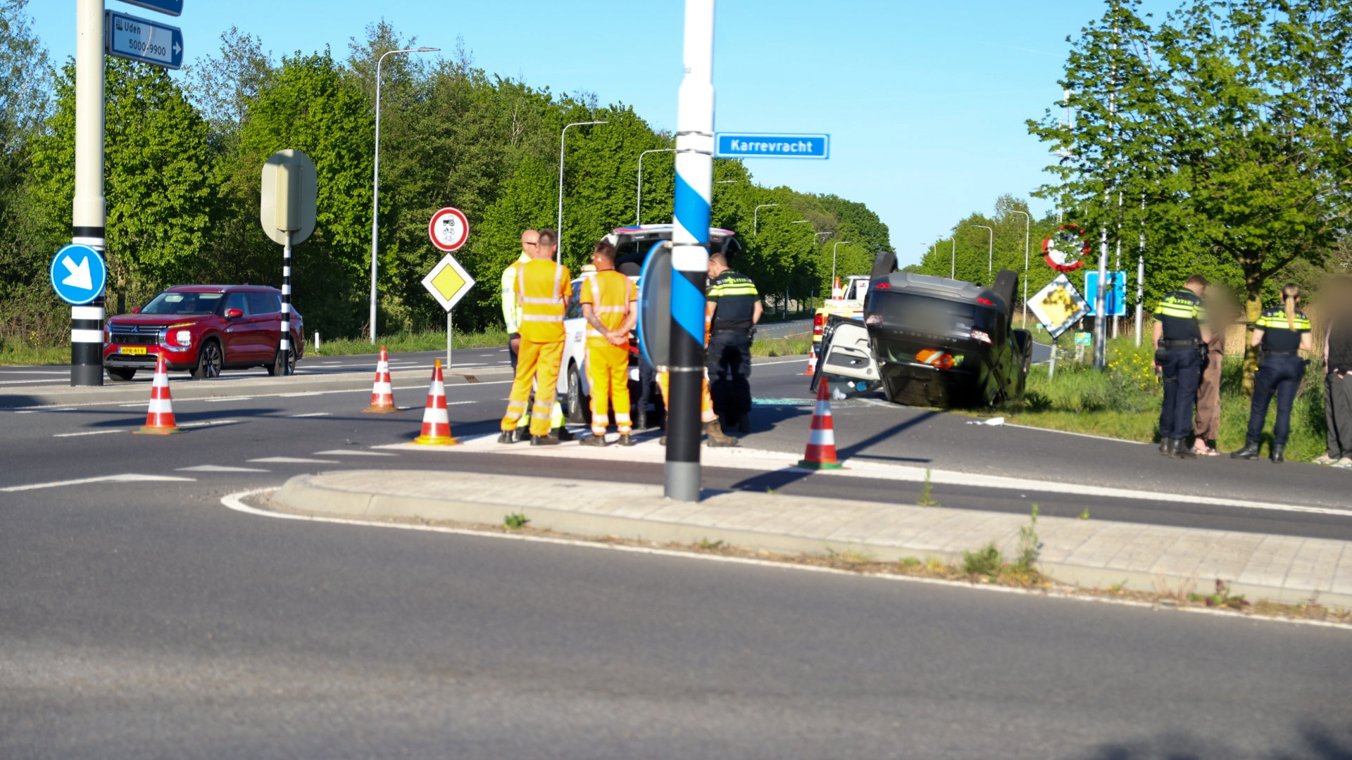 Emergency responders assess a car flipped on its roof at a road intersection, with traffic cones and bystanders nearby under clear skies.