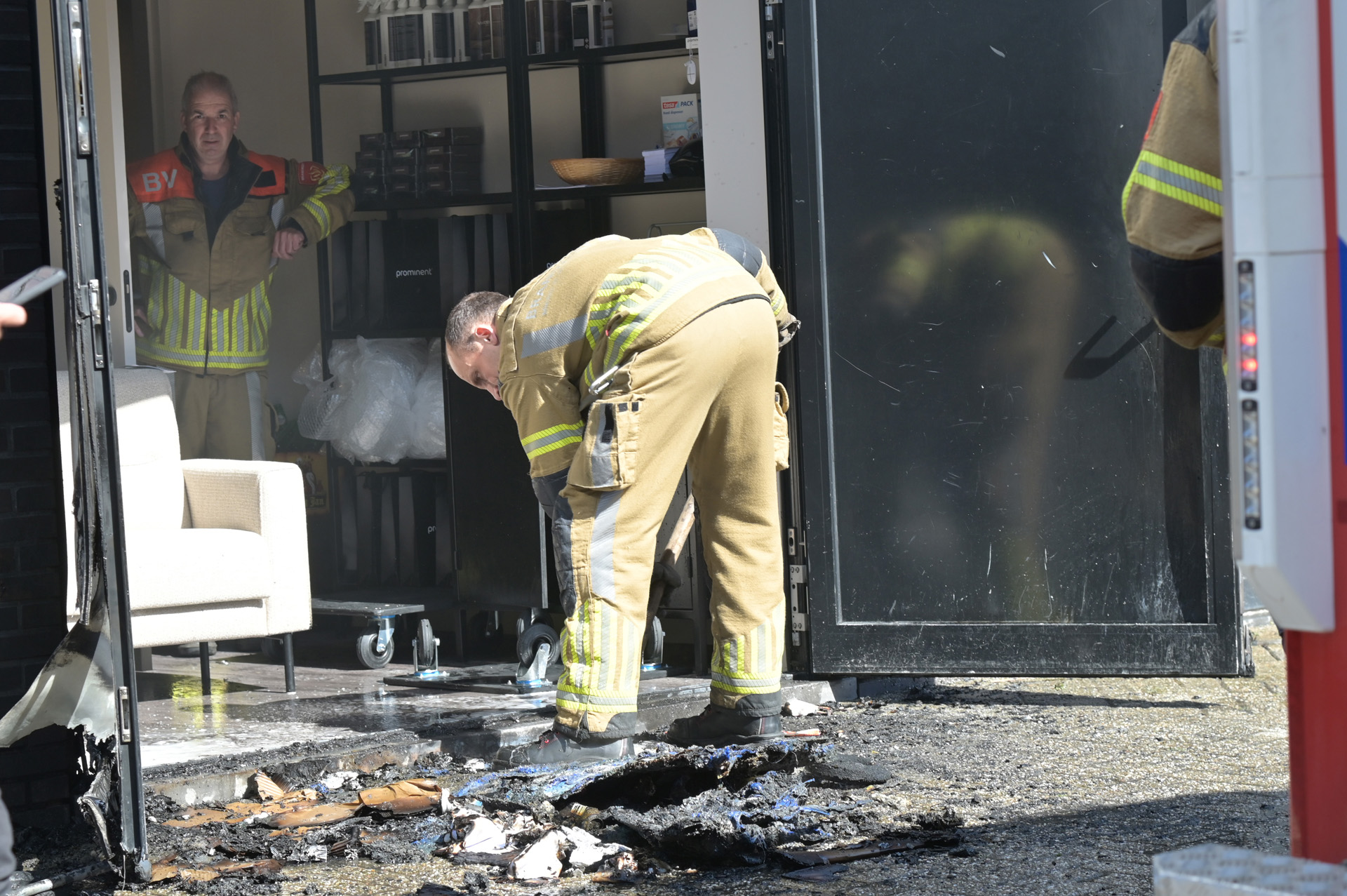 Firefighters in turnout gear stand at a smoke-damaged truck, clearing debris from the back area.