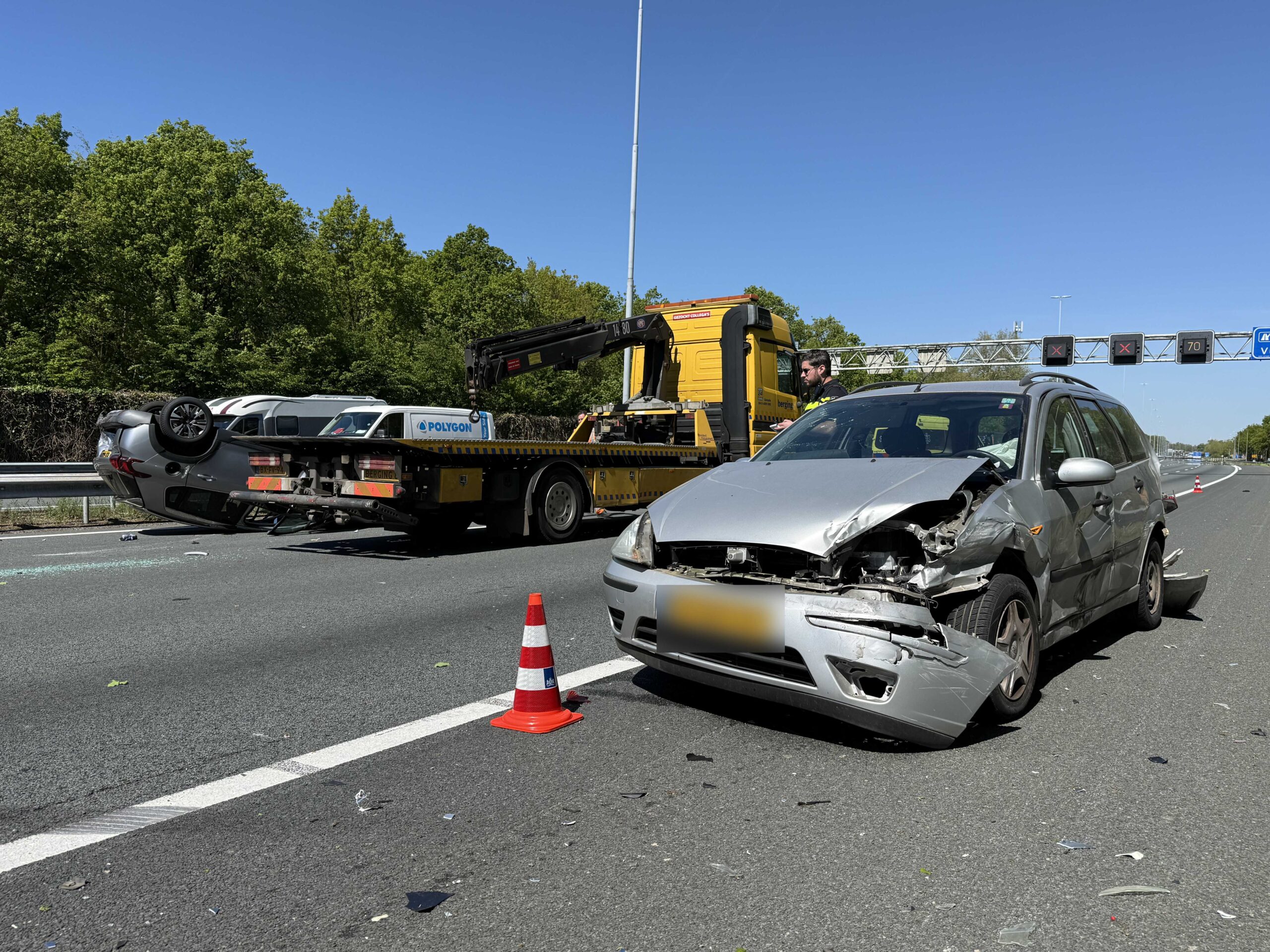 Silver car badly damaged, wedged against a tow truck on a highway shoulder under a clear blue sky; traffic cone nearby.