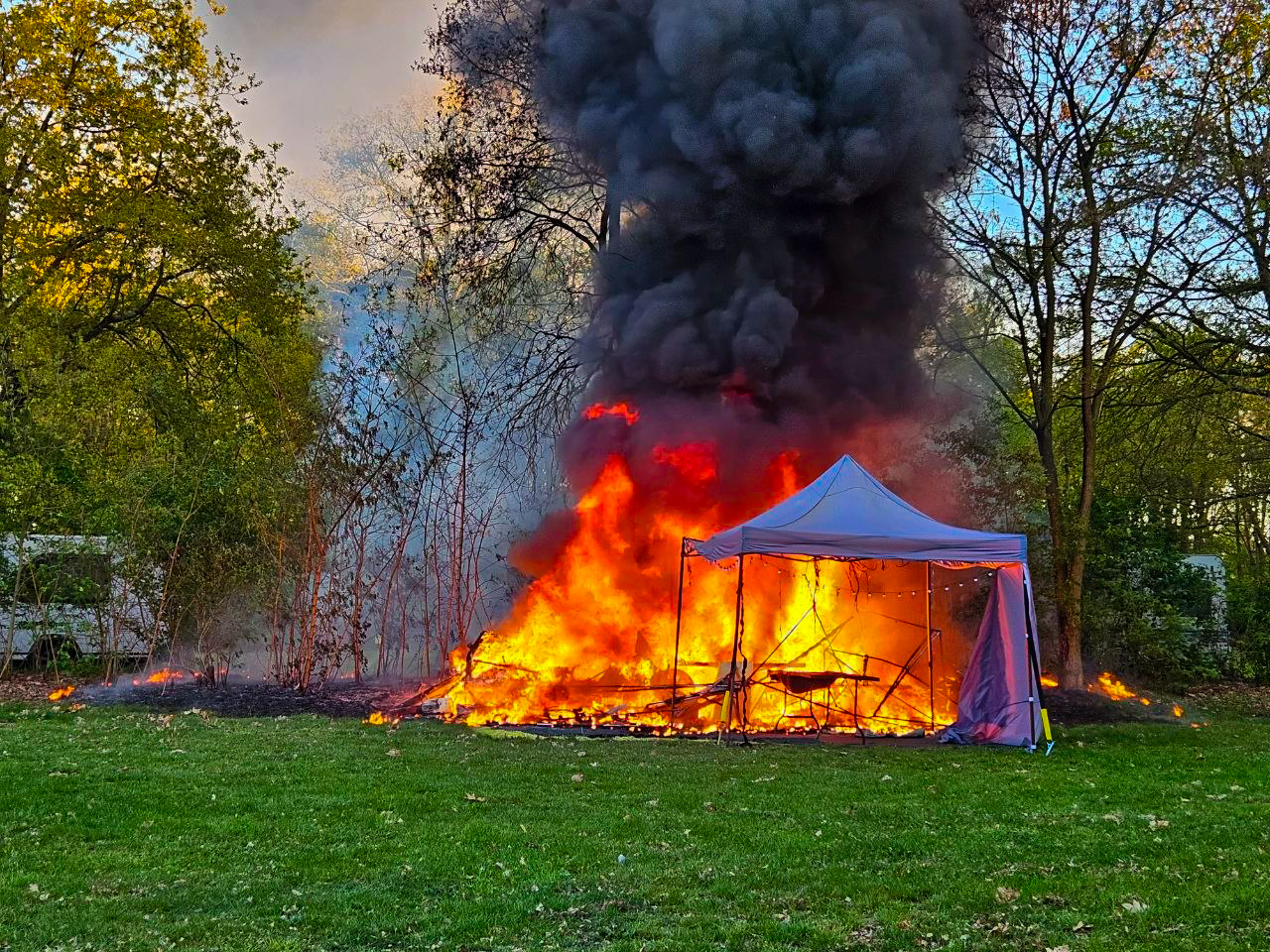 Tent on fire in a park, with thick black smoke and bright orange flames engulfing the structure.