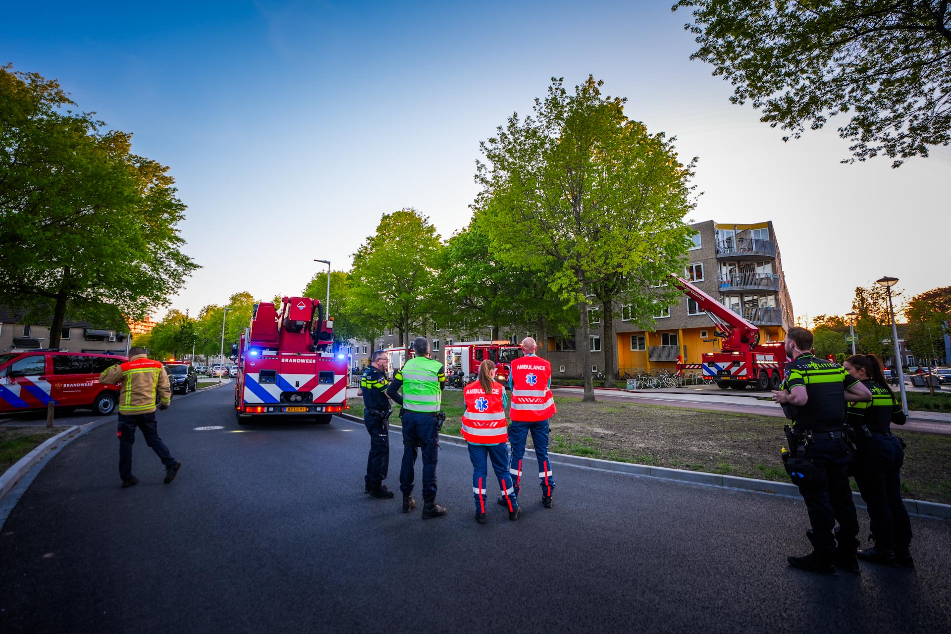 Emergency responders and red fire engines on a residential street, with a brick apartment building and green trees in the background.