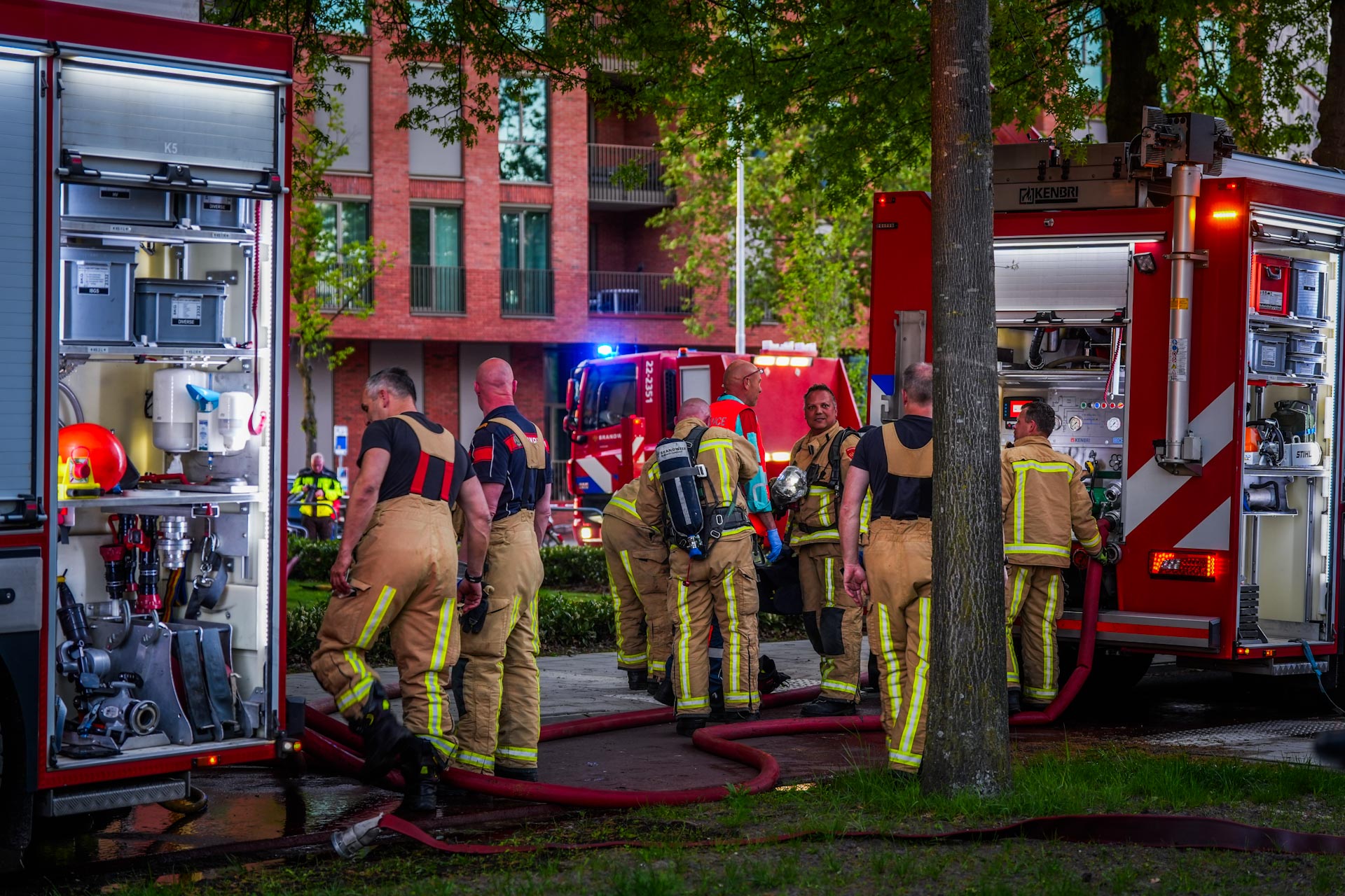 Firefighters in turnout gear gather near red fire trucks with hoses in a residential area.