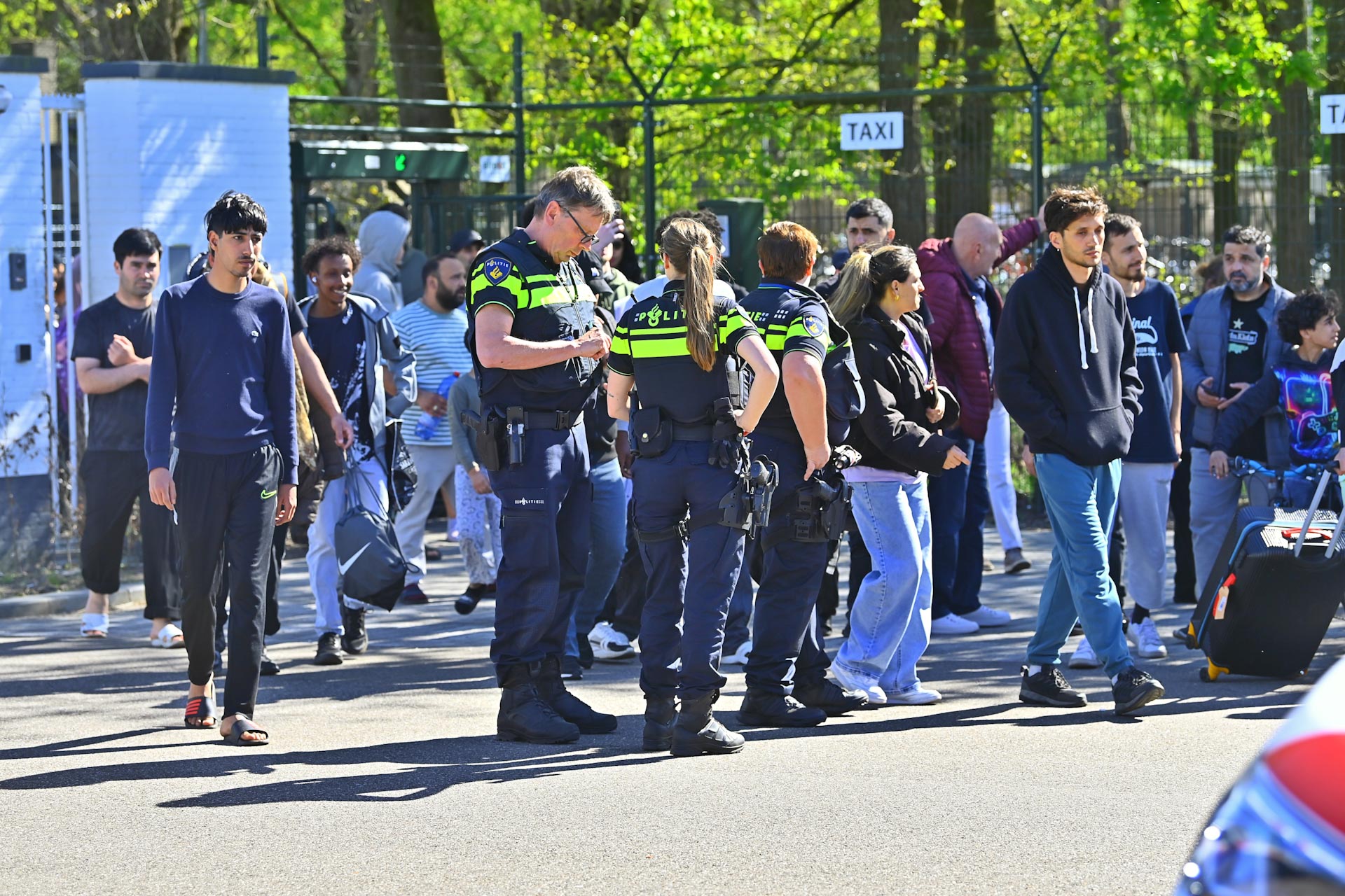 Group of people, including several police officers in high-visibility uniforms, standing and talking near a gated area on a sunny day.