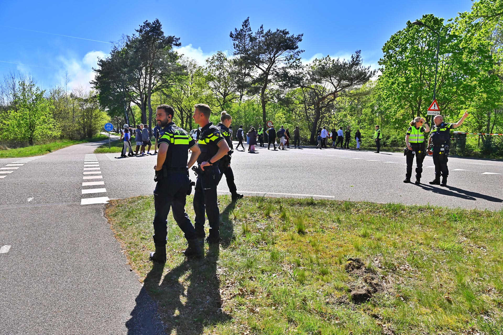Group of police officers in high-visibility vests standing on a tree-lined road with a crowd in the background on a sunny day.
