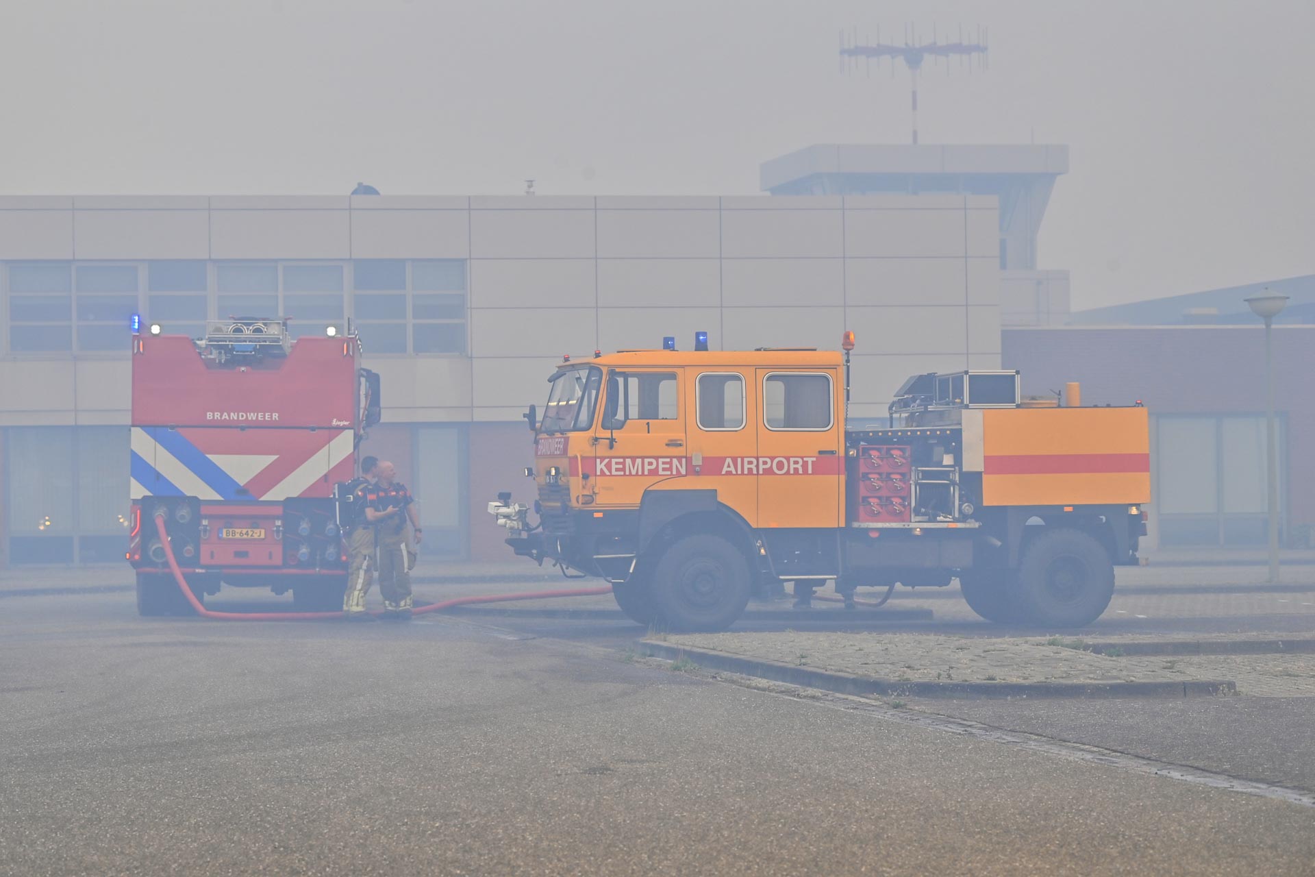 Orange airport fire truck labeled 'KEMPEN AIRPORT' on the tarmac with firefighters and a red fire engine nearby in hazy weather at a modern terminal.