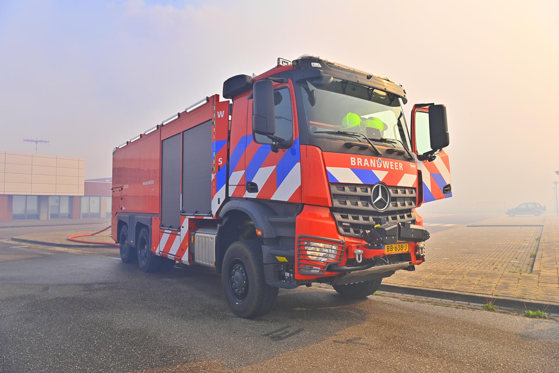 Front-right view of a red fire truck with chevron markings parked on a paved street, a hose on the ground nearby, and a pale business complex in the background.