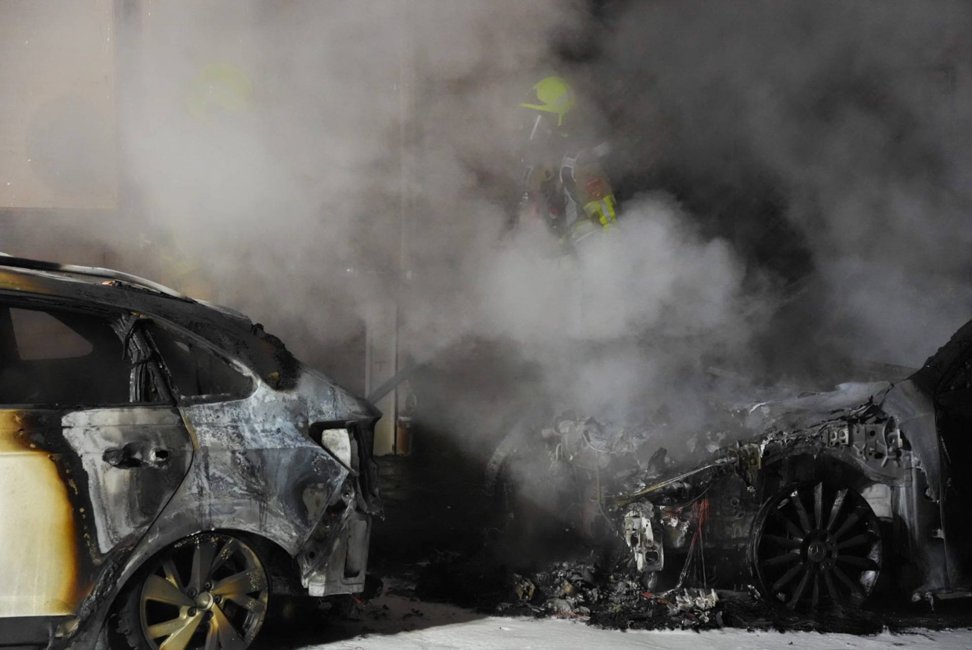 Firefighters wearing yellow helmets struggle through thick smoke among severely burned cars in a charred parking area.
