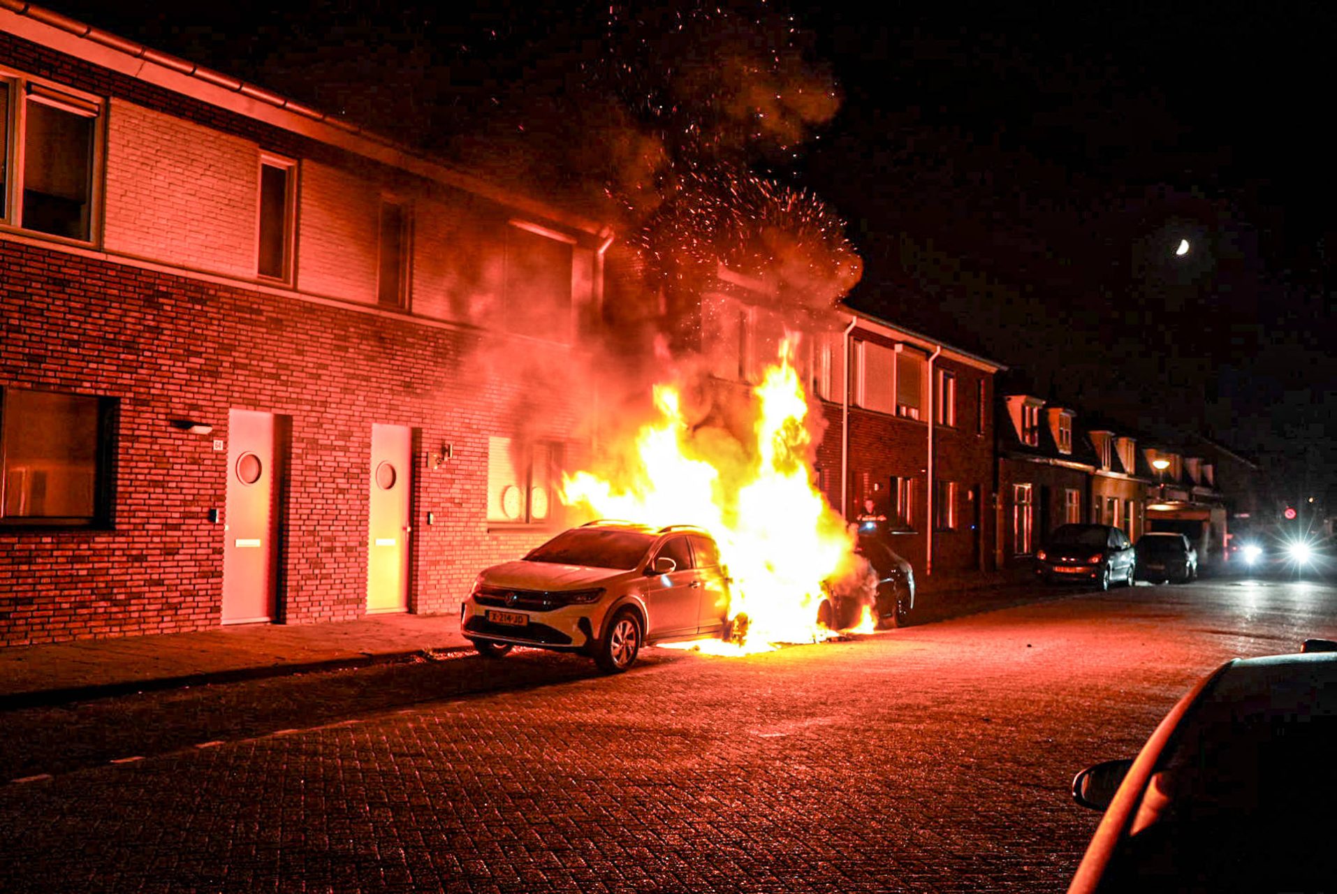 Night scene of a car on fire on a brick townhouse street, flames and smoke engulfing the vehicle.