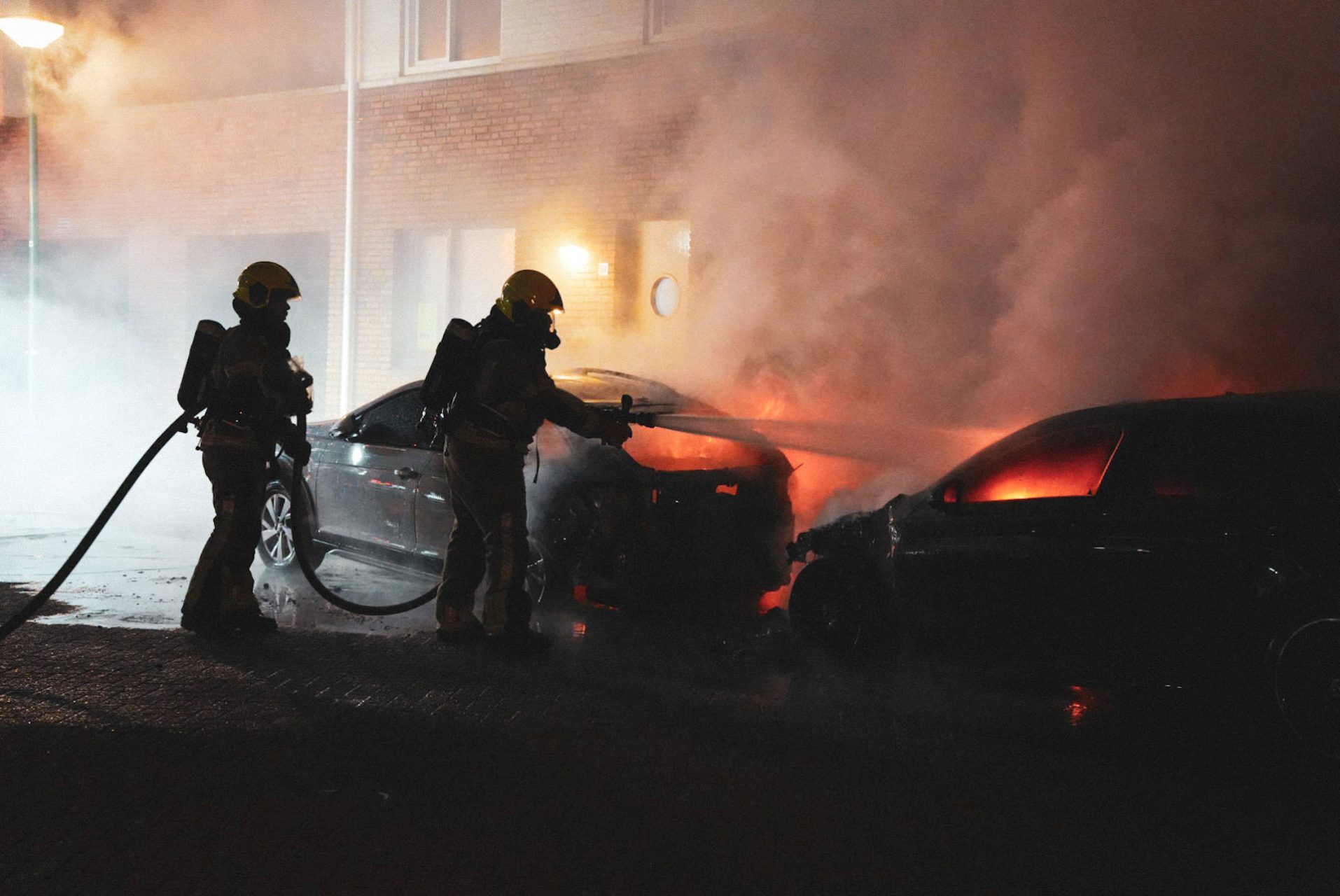 Two firefighters in turnout gear spray water on a burning car at night, smoke filling the street outside a brick building, headlights glowing.