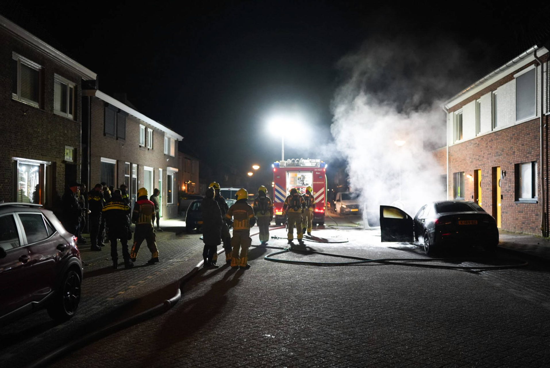 Nighttime firefighting scene on a residential street: firefighters in gear near a red fire engine with smoke rising nearby.