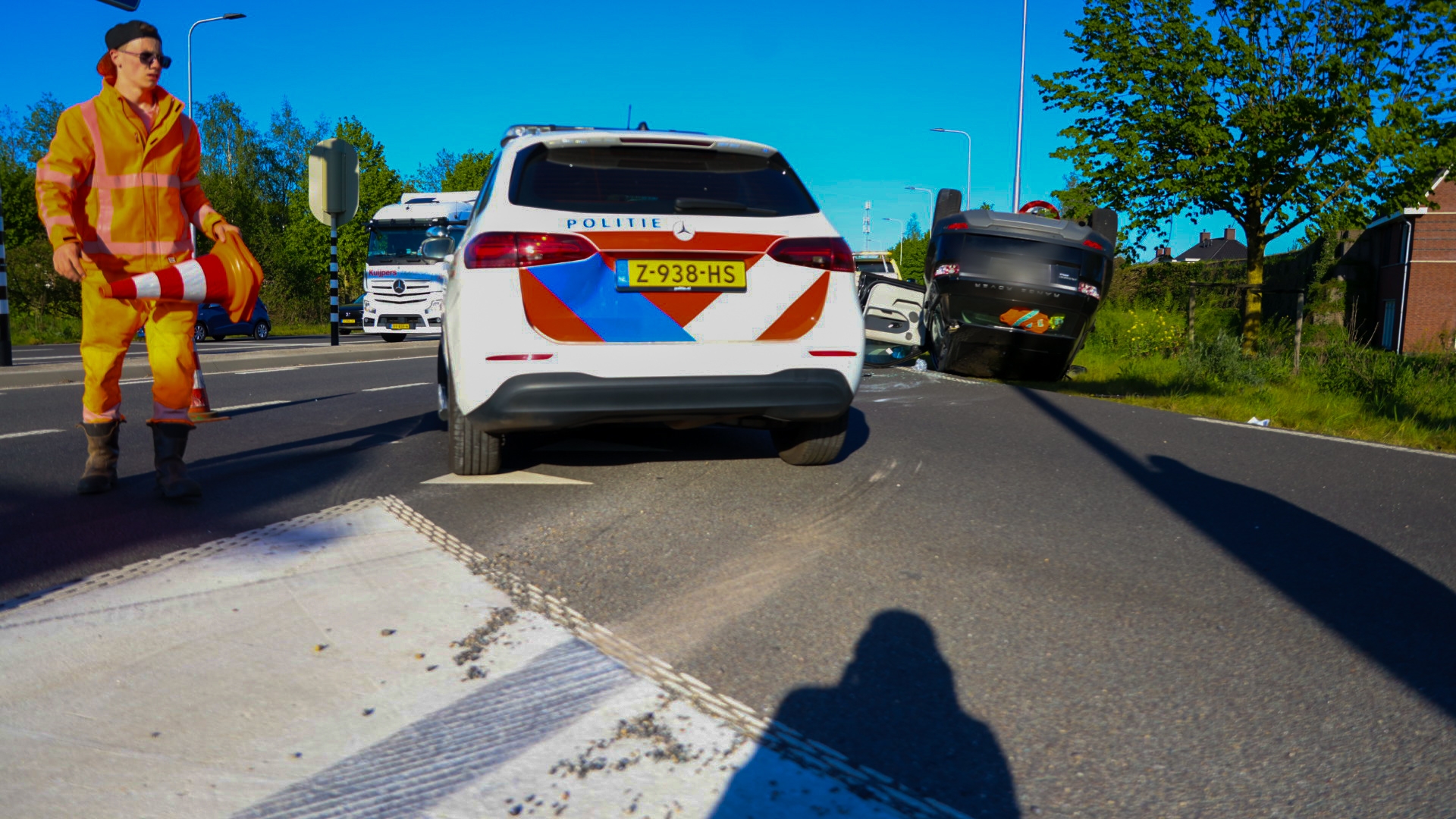 Police car with 'POLITIE' markings on a multi-vehicle crash, a black overturned car on the roadside, and a worker in an orange safety suit directing traffic.