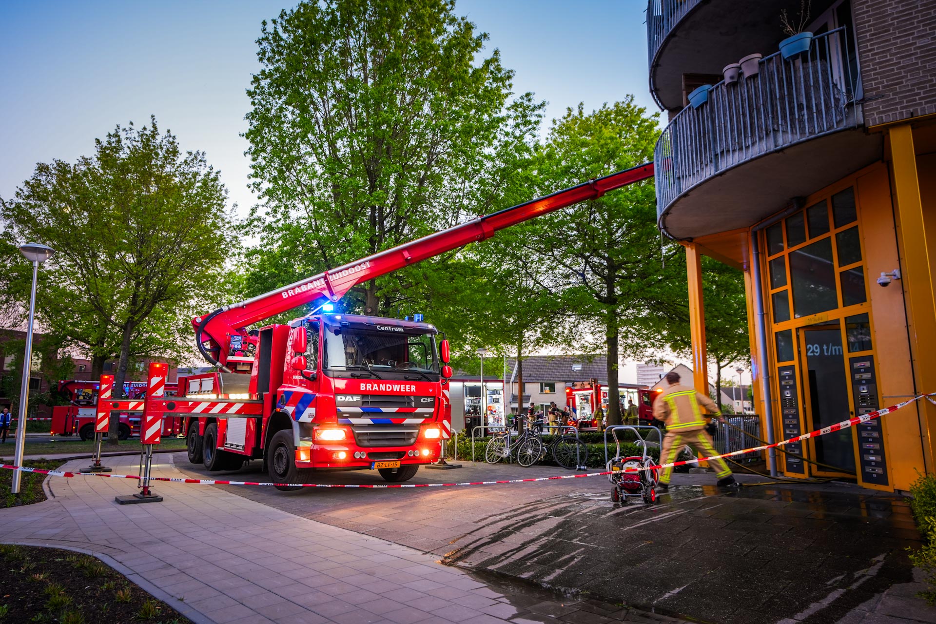 Fire trucks with an extended red ladder reaching a building, firefighters on scene in a city plaza with caution tape.