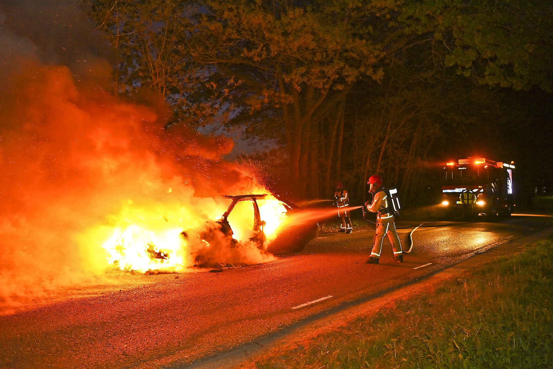 Car on fire on a road at night, firefighters spraying water while a fire truck with lights responds in the background.