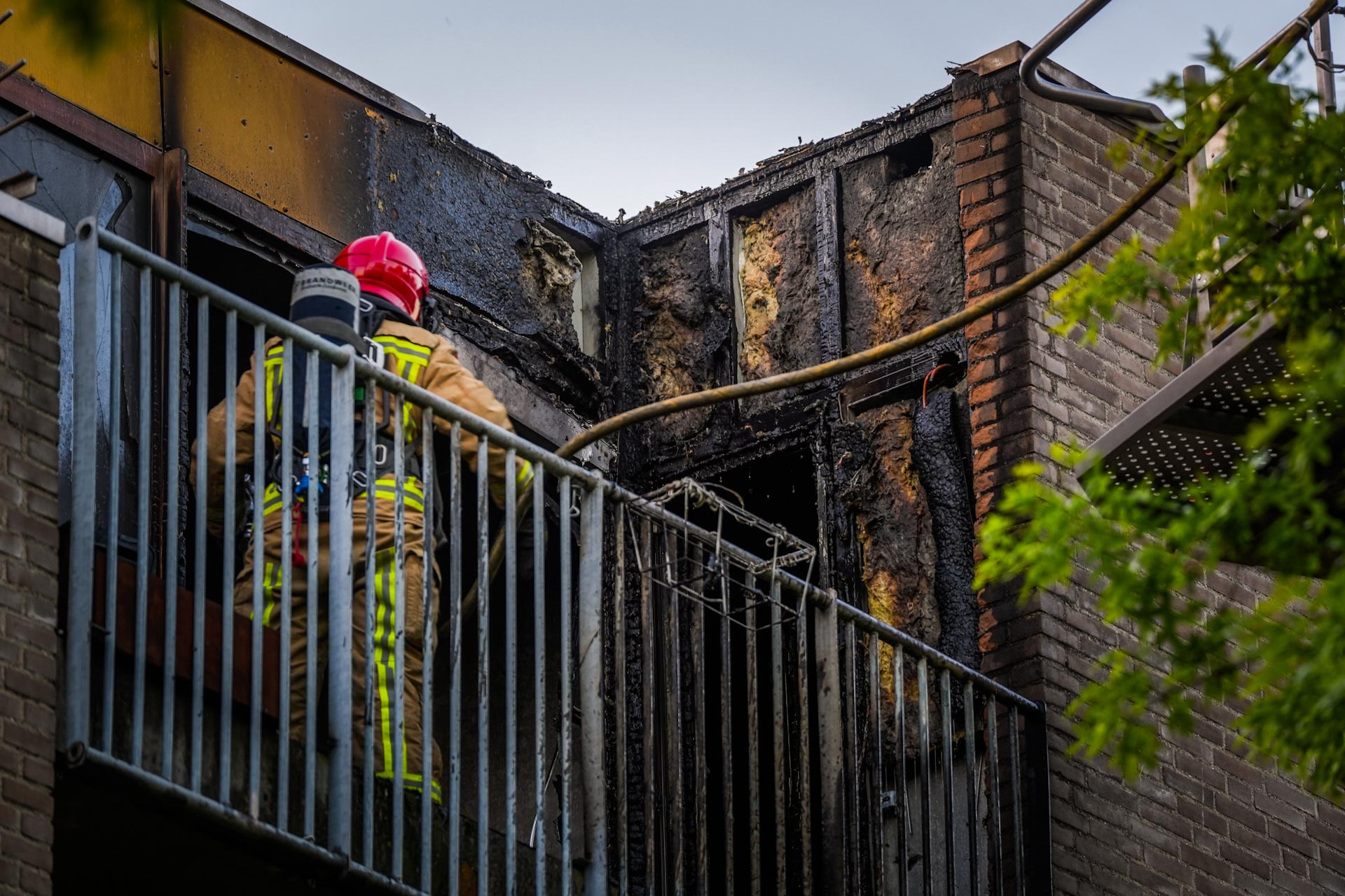 Firefighters in protective gear on a smoke-darkened balcony surveying a burned brick building after a fire.