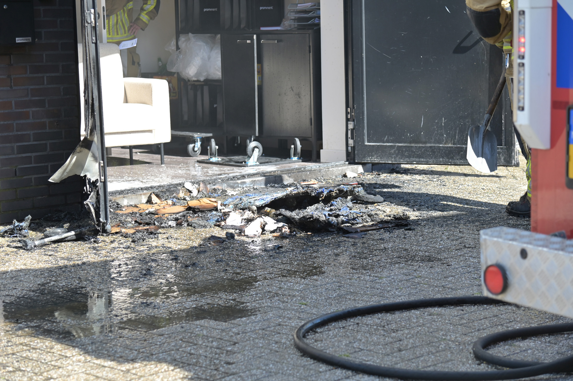 Storefront debris and charred entrance after a fire; wet ground and hoses nearby with a firefighter in gear present in the background, handling cleanup edge of scene.