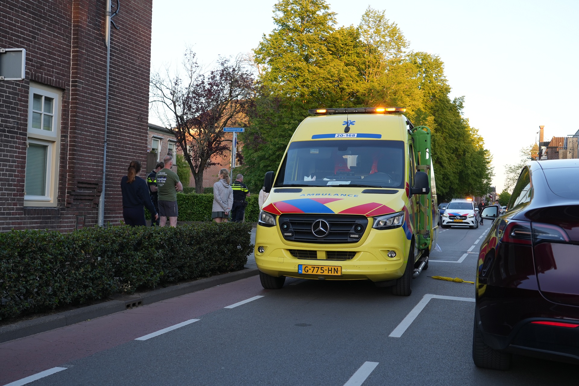 Yellow emergency ambulance parked on a residential street beside a brick building, with people standing on the sidewalk and trees in the background.