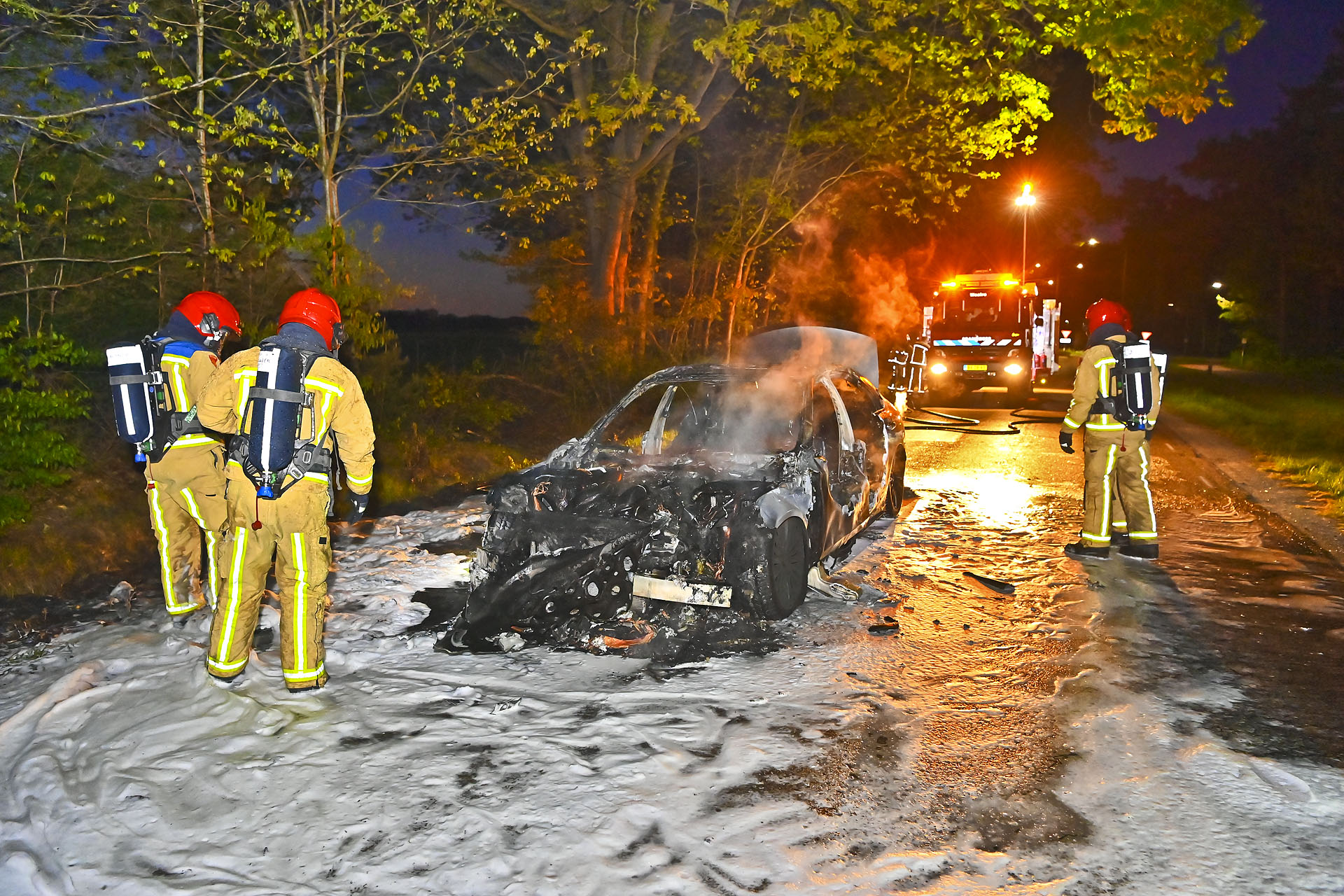 Firefighters in red helmets and turnout gear standing beside a charred car on a snowy road at night; a fire truck with lights in the background.