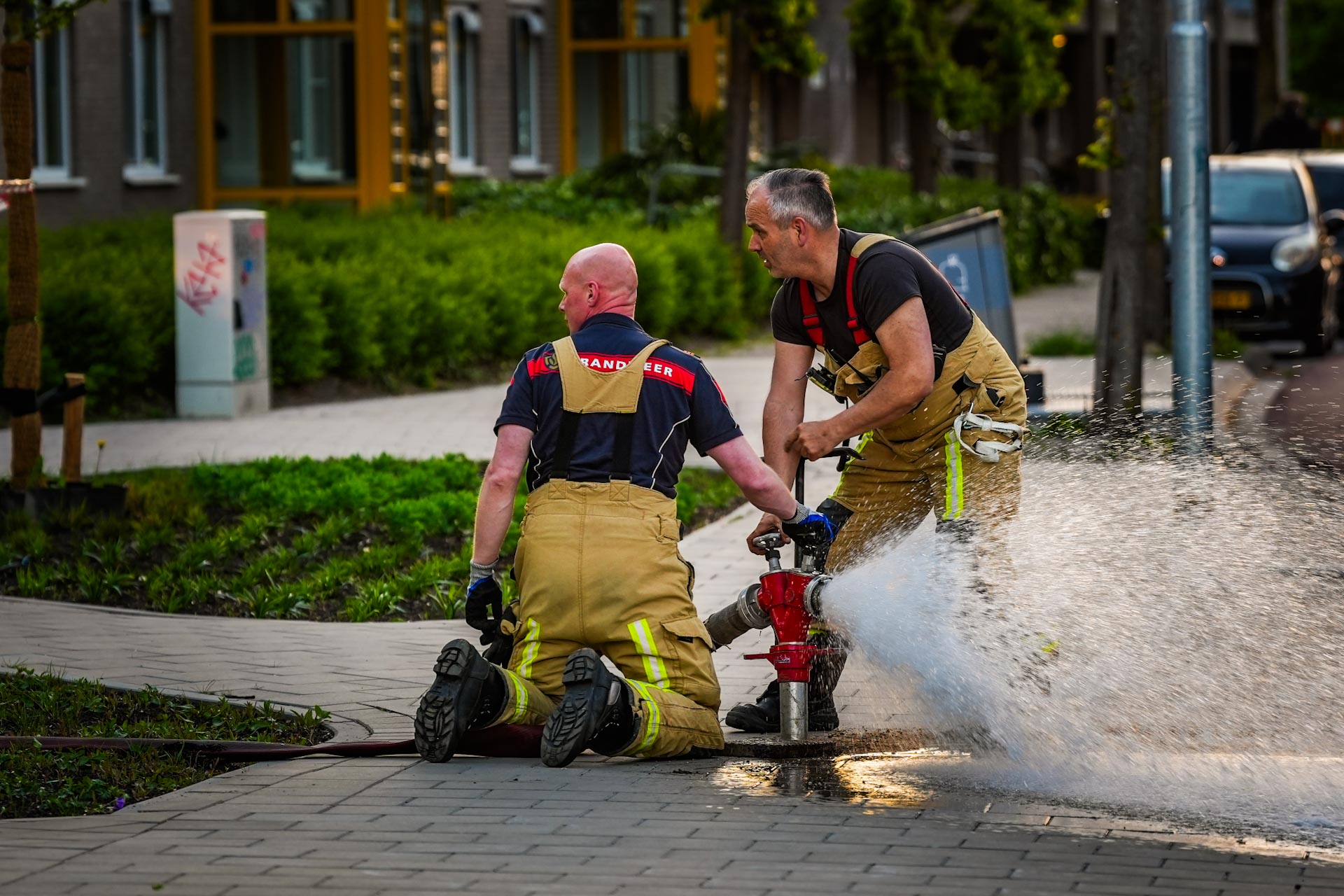 Two firefighters in tan turnout gear kneel on a wet sidewalk, directing a high-pressure hose spraying water across the scene.