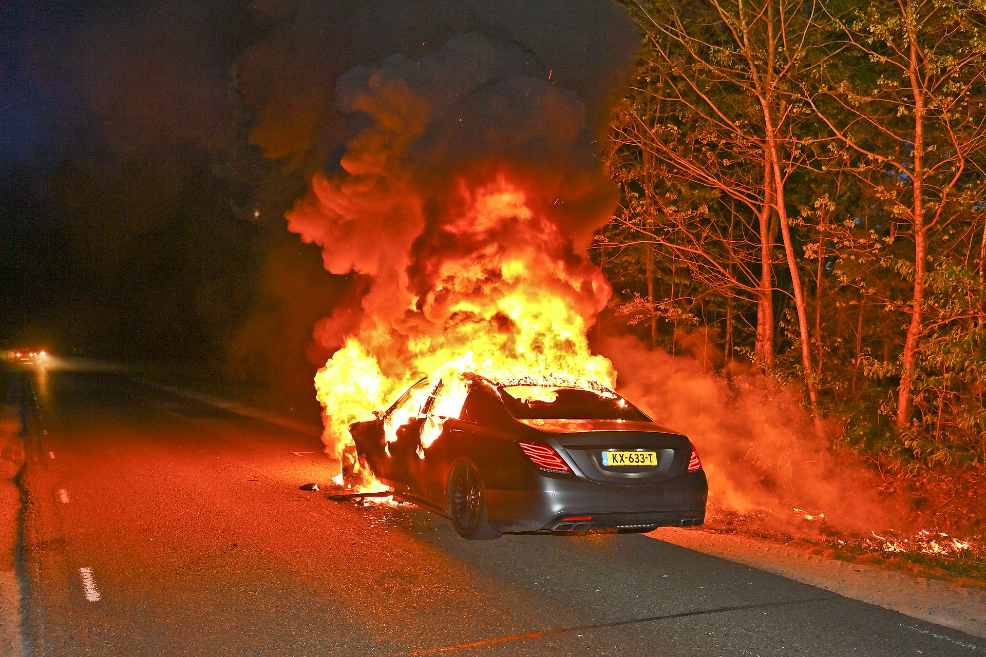 Nighttime car fire: a dark sedan on a road engulfed in bright orange flames with thick smoke and trees to the right side.