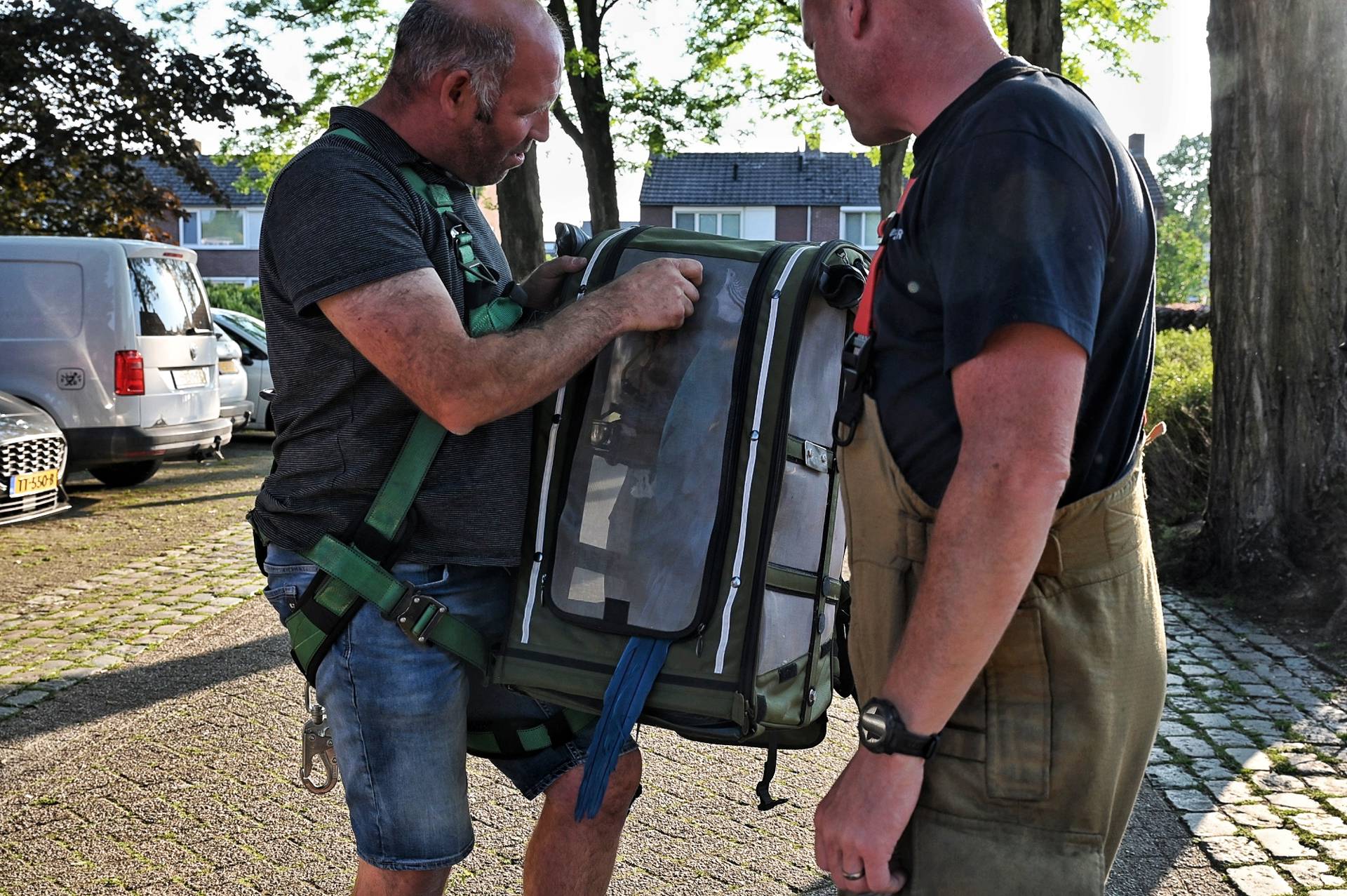 Two men wearing outdoor gear tend to a large soft-sided pet carrier resting between them outdoors.