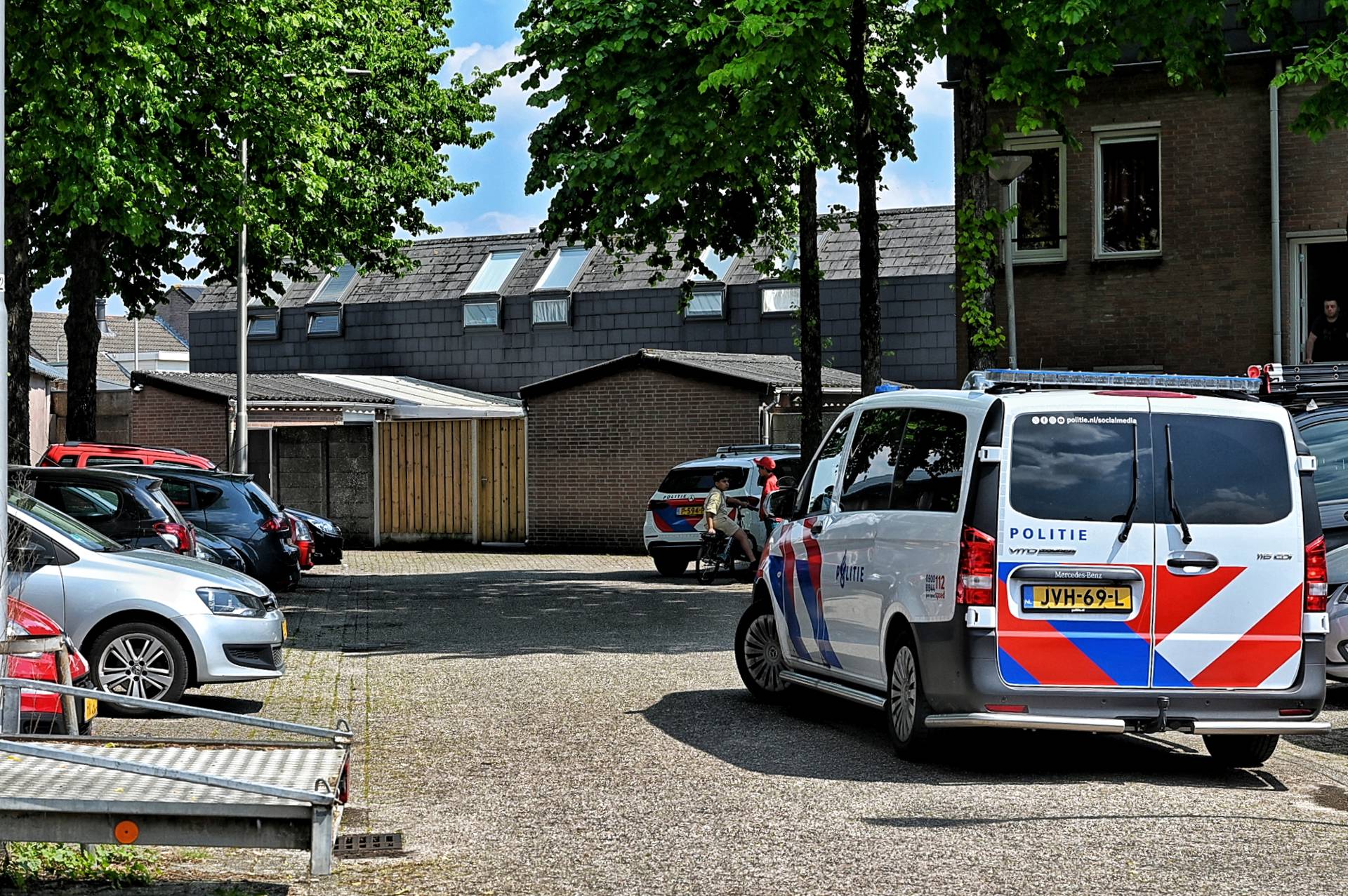 Police vans and parked cars in a residential driveway area with trees overhead and brick buildings in the background.
